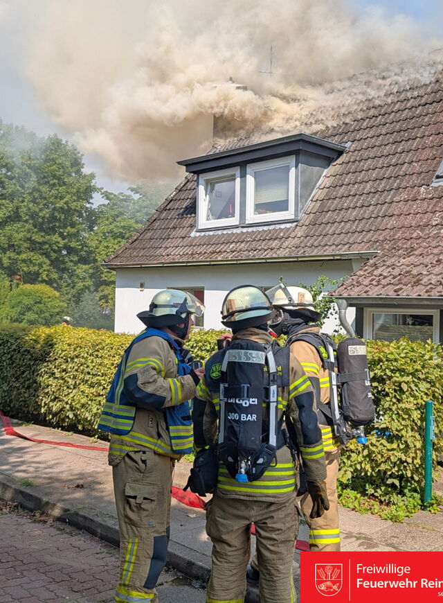 Drei Feuerwehrleute in kompletter Schutzkleidung und mit Atemschutzgeräten stehen vor einem Wohnhaus. Vom Dach des Hauses steigt dichter, heller Rauch auf. Ein roter Feuerwehrschlauch liegt auf dem Gehweg.